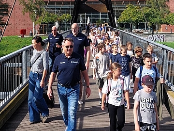 Eine Gruppe von Kindern und Erwachsenen überquert bei Sonnenschein die Fußgängerbrücke zur Insel Ziegenwerder in Frankfurt (Oder). Im Hintergrund ist ein rotes Backsteingebäude zu sehen.